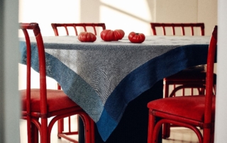 Tavolo rotondo decorato con tovaglia Society Limonta dalle sfumature blu, al centro pomodori freschi e sedie rosse tutt’intorno.